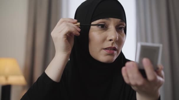 Close-up of young beautiful Muslim lady looking at small mirror and applying mascara. Modern eastern woman in black hijab doing make-up. Beauty, cosmetics. - Imágenes, Vídeo