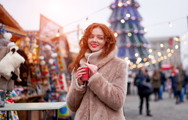 Young beautiful redhead girl freckles ice rink on background. Pretty woman curly hair portrait walking on new year fair - Fotó, kép