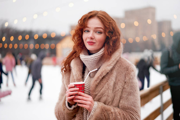 Young beautiful redhead girl freckles ice rink on background. Pretty woman curly hair portrait walking on new year fair - Foto, Imagen