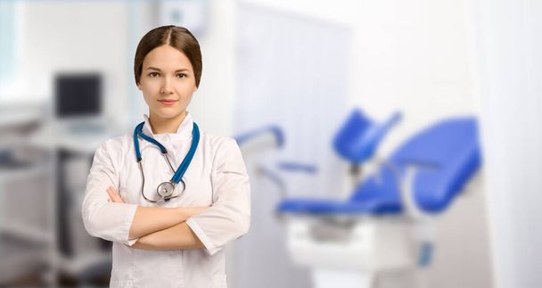 Girl doctor gynecologist in a white medical gown with a stethoscope stands in Cabinet against background of gynecological equipment and looks at camera. Place for text and design. - Photo, Image