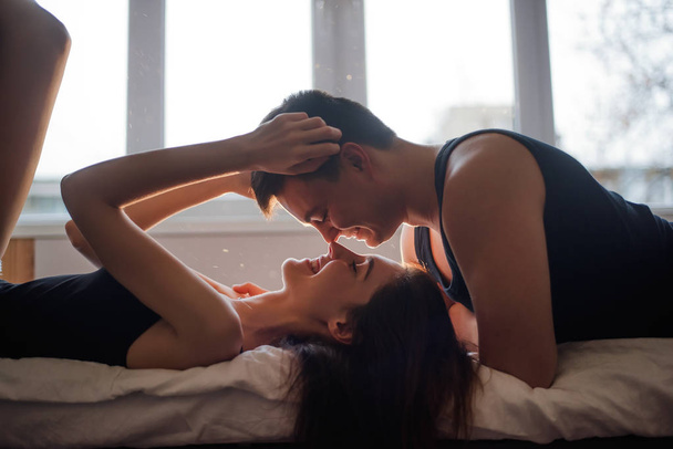 The concept of tactility, trust in a partner. A couple in love lies on the bed, legs up, laughing, hugging at home. A young man tenderly kisses a woman at the window of loft. Lovers hands touch. - Fotografie, Obrázek
