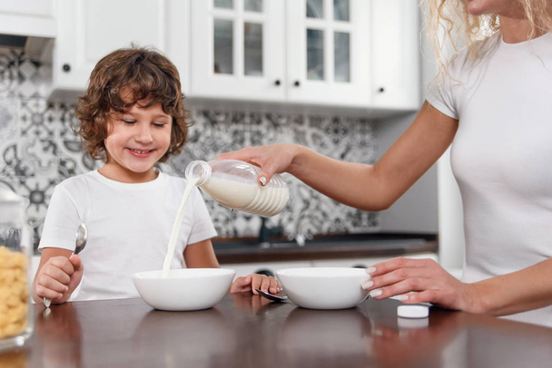 Beautiful mother pours milk into a plate with cereal Beautiful mother prepares breakfast for her beloved son - Φωτογραφία, εικόνα