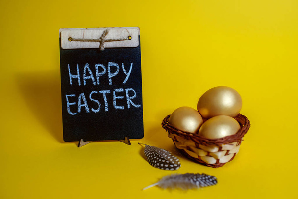La inscripción Feliz Pascua en la pizarra, huevos de gallina dorados sobre un fondo amarillo en una canasta de mimbre, plumas de codorniz
 - Foto, imagen