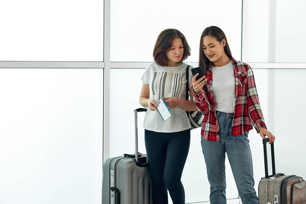 Two girls using smartphone checking flight or online check-in at airport, with luggage. Air travel, summer holiday, or mobile phone application technology concept - Photo, image