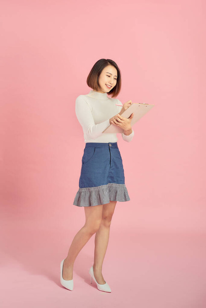 Full length of a smiling Asian woman standing isolated over pink background, holding notepad and pencil - Фото, зображення