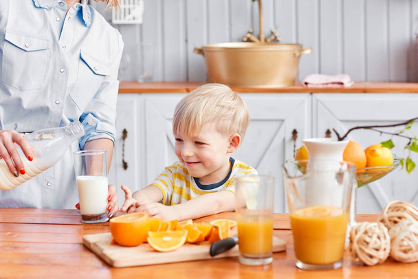 Moeder en zoon zijn glimlachen terwijl het hebben van een ontbijt in de keuken. Moeder is het gieten van melk in glazen - Foto, afbeelding
