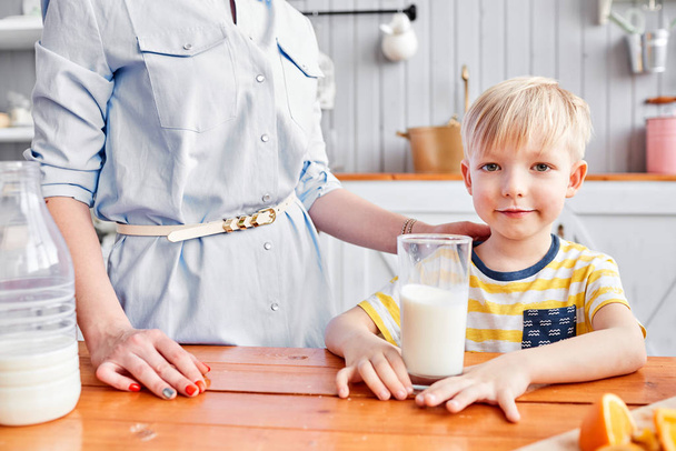 Küçük çocuk mutfakta kahvaltı ederken gülümsüyor. Annem bardağa süt döküyor. - Fotoğraf, Görsel