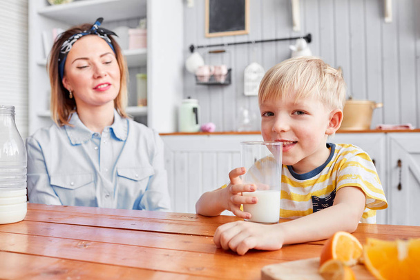 Madre e hijo sonríen mientras desayunan en la cocina. Mañana brillante en la cocina. Desayuno saludable cereales y fruta fresca
. - Foto, imagen