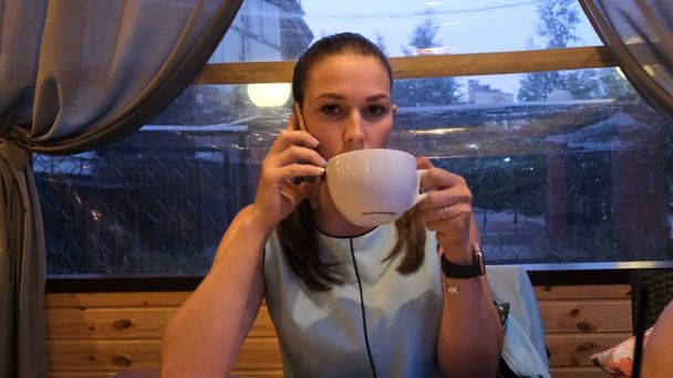 Жінка в блакитній сукні п'є каву і розмовляє по телефону в кафе ввечері
 - Фото, зображення