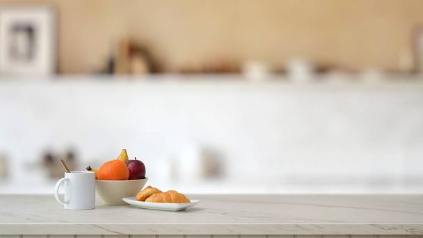 Vista de cerca de frutero, taza de café y croissant en escritorio de mármol con sala de cocina borrosa
  - Foto, imagen