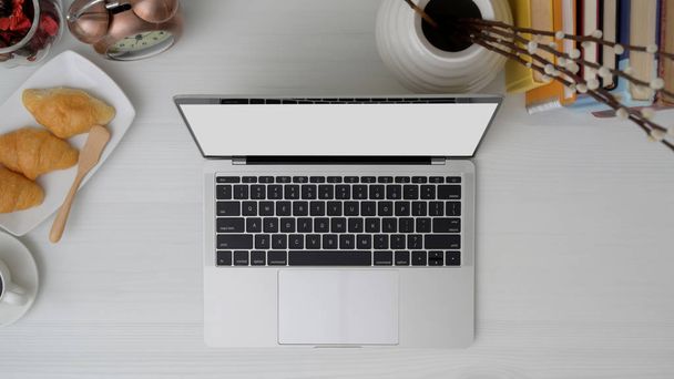 Irodai helyiség feletti felvétel laptoppal, kávéfőzővel, croissant-nal és irodai kellékekkel fehér asztalon  - Fotó, kép