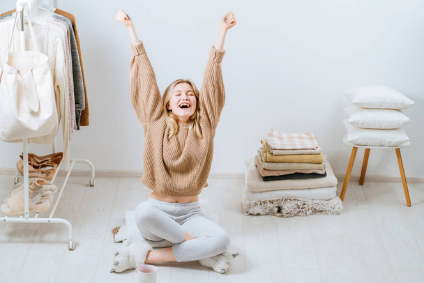 Young woman sitting on the floor near stack of towels pillows, cover plaid on white wall stack tower next to her on chair. Home in scandinavian style, clothes rack in modern empty wardrobe room. - Φωτογραφία, εικόνα