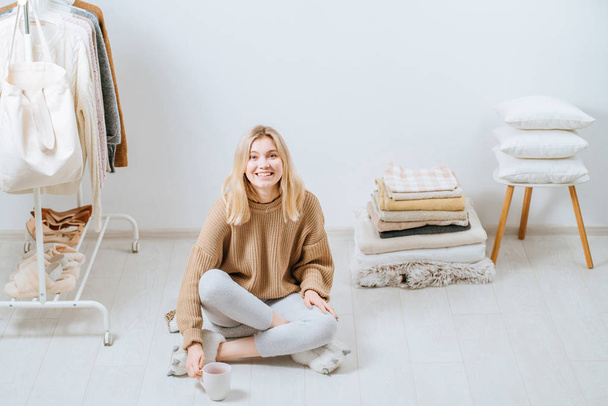 Young sad thoughtful woman sitting on the floor drinking coffee around morning routine near stack of towels pillows, plaid. Home in scandinavian style, clothes rack in modern empty wardrobe room. - Foto, imagen