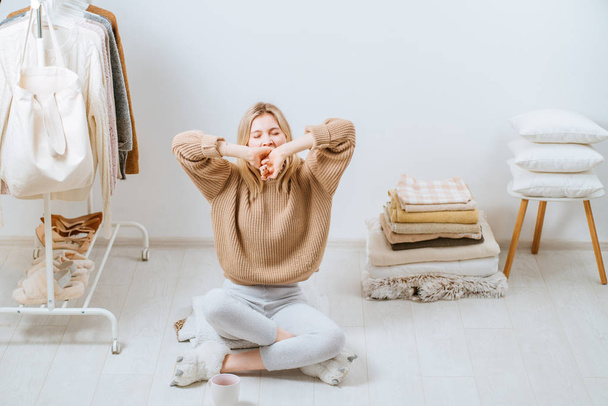 Young woman sitting on the floor near stack of towels pillows, cover plaid on white wall stack tower next to her on chair. Home in scandinavian style, clothes rack in modern empty wardrobe room. - Foto, Imagem