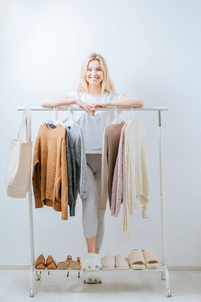Young pretty blond girl blogger standing, having difficult choice not knowing what to put on on the background of clothes hanging on a hanger in the wardrobe. Full hoight photo - Foto, imagen