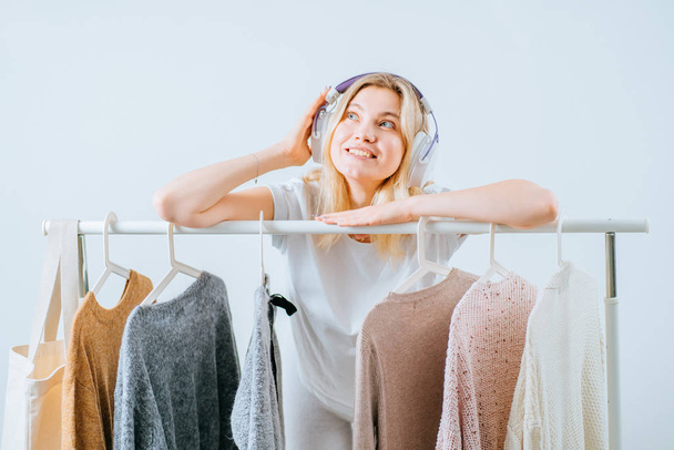 Beautiful young friendly positive woman blogger influencer near rack with hangers on white background. Concept choice not knowing what to put on. - Photo, Image