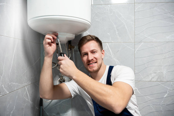 młody przystojny uśmiechnięty mężczyzna pracownik w mundurze ze śrubokrętem w ręku naprawy podgrzewacza wody w domu w toalecie profesjonalny serwis naprawczy - Zdjęcie, obraz