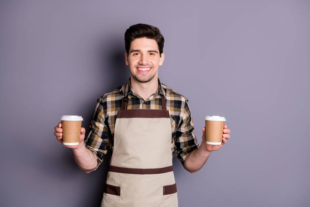 Photo of attractive worker guy holding hands two paper coffee cups inviting visitors try aroma coffee professional barista wear apron plaid shirt isolated grey color background - Fotografie, Obrázek