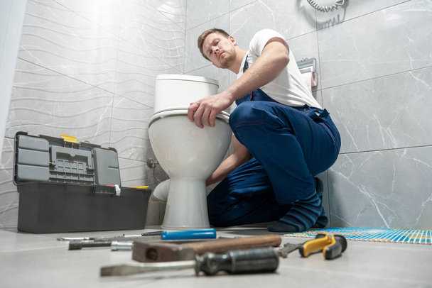 man plumber in uniform repairing toilet bowl using instrument kit professional repair service - Photo, Image