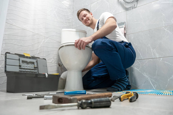 smiling man plumber in uniform repairing toilet bowl using instrument kit looks happy professional repair service - Zdjęcie, obraz