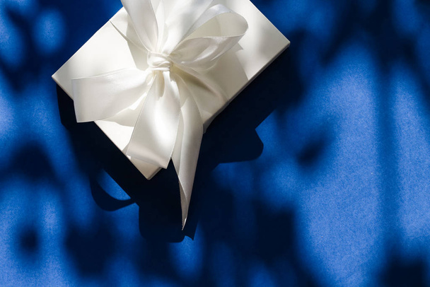 Розкішна святкова біла подарункова коробка з шовковою стрічкою та бантом на синьому б
 - Фото, зображення