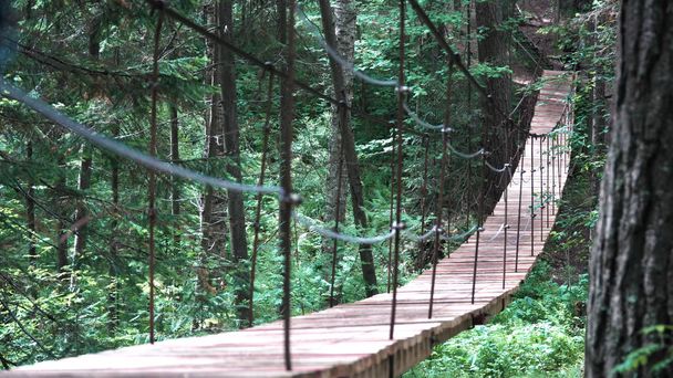 A suspension bridge on hiking trail through green dense forest with a man traveler with red backpack. Stock footage. Rear view of a man crossing the hanging bridge. - Foto, Bild