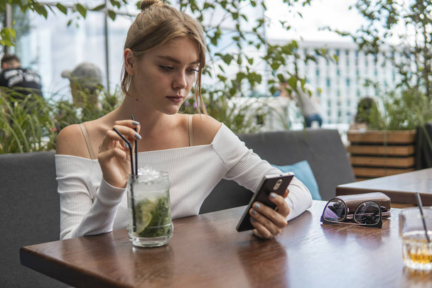 カクテルと陽気なブロンドの女性ブロガーは、カフェで無料のwifiに接続されているスマートフォンで着信SMSメッセージを読んでいます。現代の携帯電話でオンラインで放送を見て肯定的な若い学生. - 写真・画像