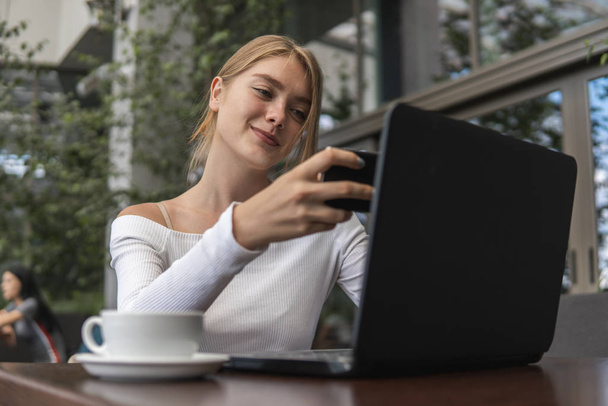 Moderne zakenvrouw leest inkomende sms-berichten op smartphone of kijkt online naar uitzendingen op moderne mobiele telefoon terwijl ze met een laptop in de caffe zit. Vrouwelijke student zit in koffie. - Foto, afbeelding