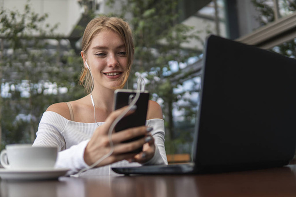 Modern business woman is reading incoming sms message on smartphone or watching broadcasting online on modern mobile phone while sitting in caffe with a laptop. Female student sitting in coffee. - Foto, Bild