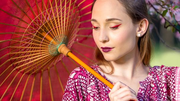 Japanese woman in kimono and with umbrella. Beauty Asian girl with a bright make-up art in green scenery park - Fotografie, Obrázek