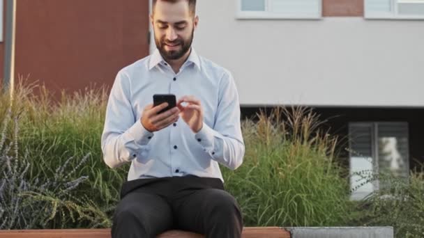 Joyful bearded businessman using smartphone while sitting on bench outdoors - Кадры, видео