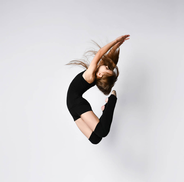 Young girl gymnast in black sport body and uppers jumping and making dymnastic pose in air over white background - Photo, Image