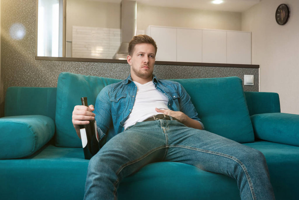 young handsome man sitting on the sofa with a bottle of wine drinking in the cozy living room home relaxed atmosphere - Foto, imagen