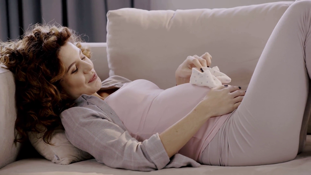 mujer embarazada feliz acostada en el sofá con zapatos de bebé
 - Imágenes, Vídeo