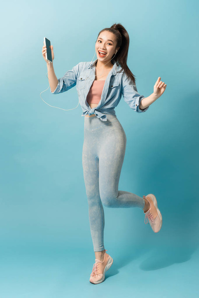 Joyful satisfeito asiático mulher em fones de ouvido ouvindo música e pulando sobre fundo azul
 - Foto, Imagem