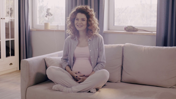 mulher grávida feliz acariciando barriga no sofá em pose de lótus
 - Filmagem, Vídeo