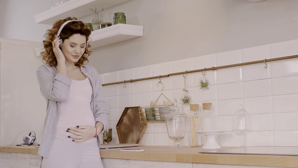 boldog terhes nő használ okostelefon és zenét hallgat a konyhában - Felvétel, videó