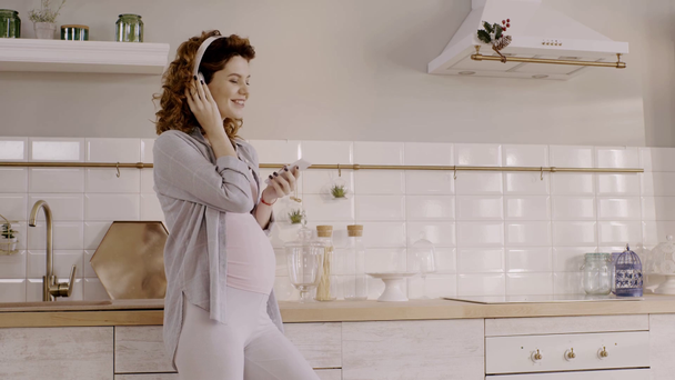 счастливая беременная женщина с помощью смартфона и прослушивания музыки на кухне
 - Кадры, видео