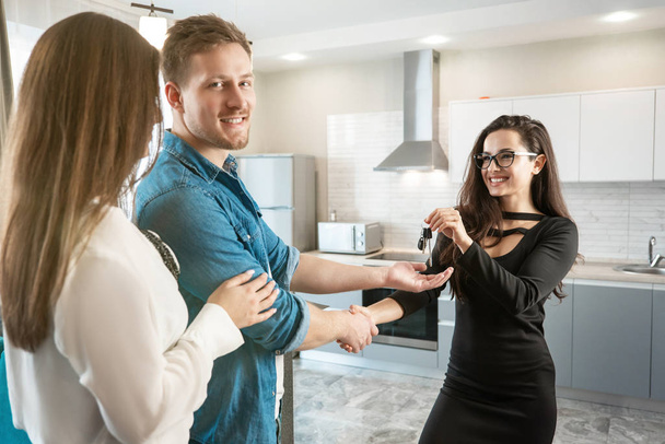 Брюнетка женщина риэлтор менеджер недвижимости давая ключи очаровательным довольным клиентам пару мужчин и женщин пожимая руку в уютных квартирах после успешного подписания договора аренды
 - Фото, изображение