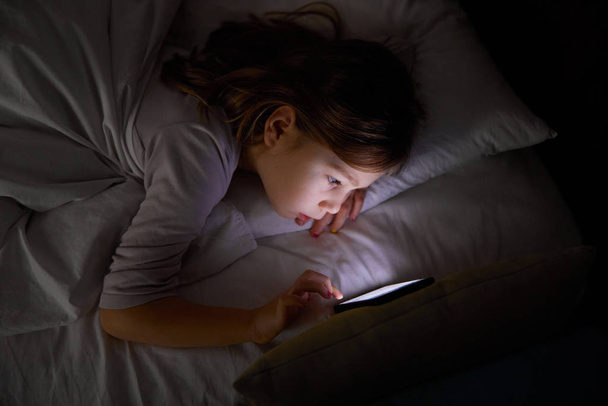 sześcioletnia dziewczyna leżąca na łóżku w nocy dotykając ekranu telefonu komórkowego - Zdjęcie, obraz