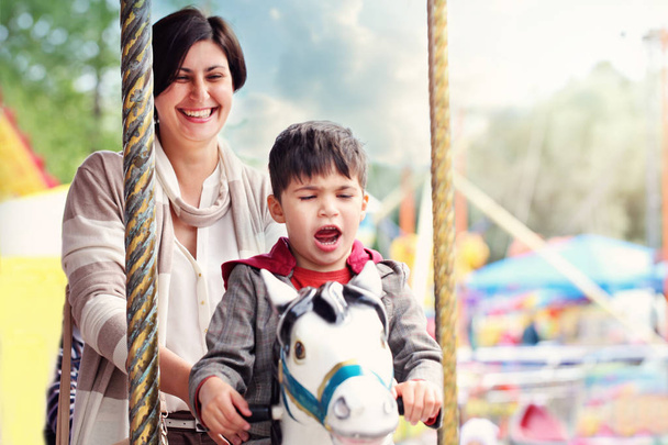Cute little boy sitting on carousel horse. His mother standing n - Foto, Imagen
