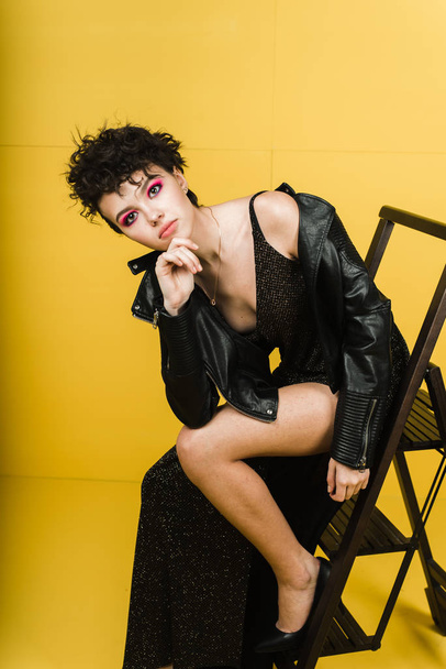 Fashionable portrait of a young girl in a leather jacket and a shiny long dress. Stylish girl in a black rock image on a yellow background in the studio - Фото, зображення