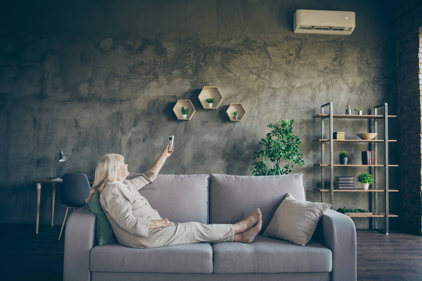Profile photo of charming white haired aged granny changing conditioner mode turn on hot air sitting sofa divan wear beige pastel clothes living room indoors - Valokuva, kuva