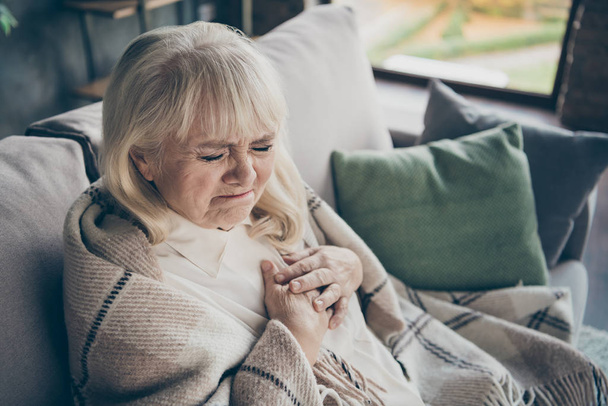 Photo of unhappy white haired aged granny holding chest zone cardiac difficulties afraid heart attack sitting sofa divan covered plaid blanket living room indoors - Фото, зображення