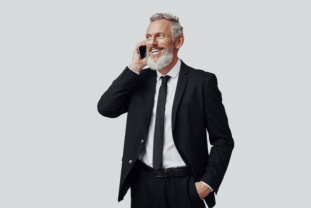 Feliz hombre maduro en traje completo hablando por teléfono y sonriendo mientras está de pie sobre fondo gris
 - Foto, imagen