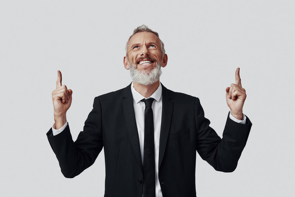 Happy mature man in full suit pointing copy space and smiling while standing against grey background - Foto, imagen