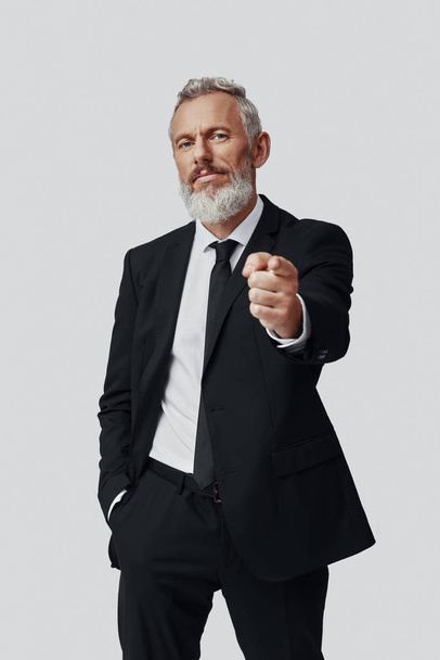 Confident mature man in full suit pointing you and looking at camera while standing against grey background - Foto, immagini