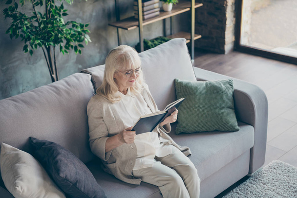 Photo of blond adorable aged granny sitting comfy sofa divan reading favorite historic novel book nice free time wear beige pastel clothes flat living room indoors - Zdjęcie, obraz