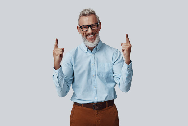 Cheerful mature man smiling and pointing copy space while standing against grey background - Zdjęcie, obraz