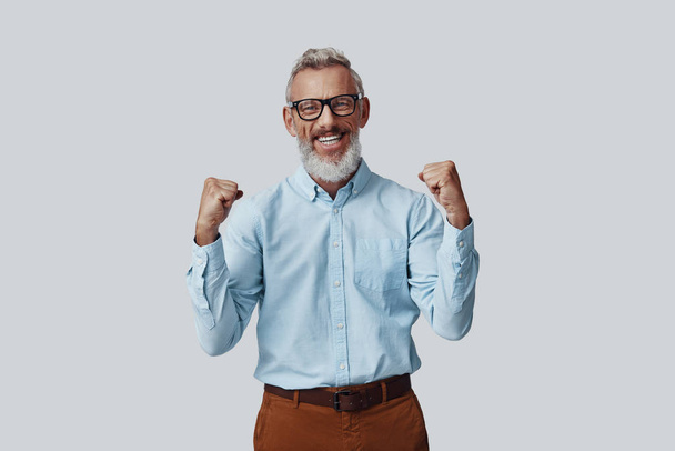 Cheerful mature man looking at camera with smile and gesturing while standing against grey background - Foto, Imagem